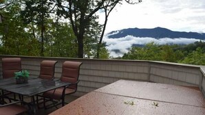 Een terras buiten met een tafel en stoelen, omringd door een lage muur, met uitzicht op een bergachtig landschap met wolken.