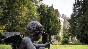 A statue of a figure with wings and a trumpet, set in a grassy area with trees and a building in the background. - Château-l'evêque en Périgord (Château-l'Évêque)