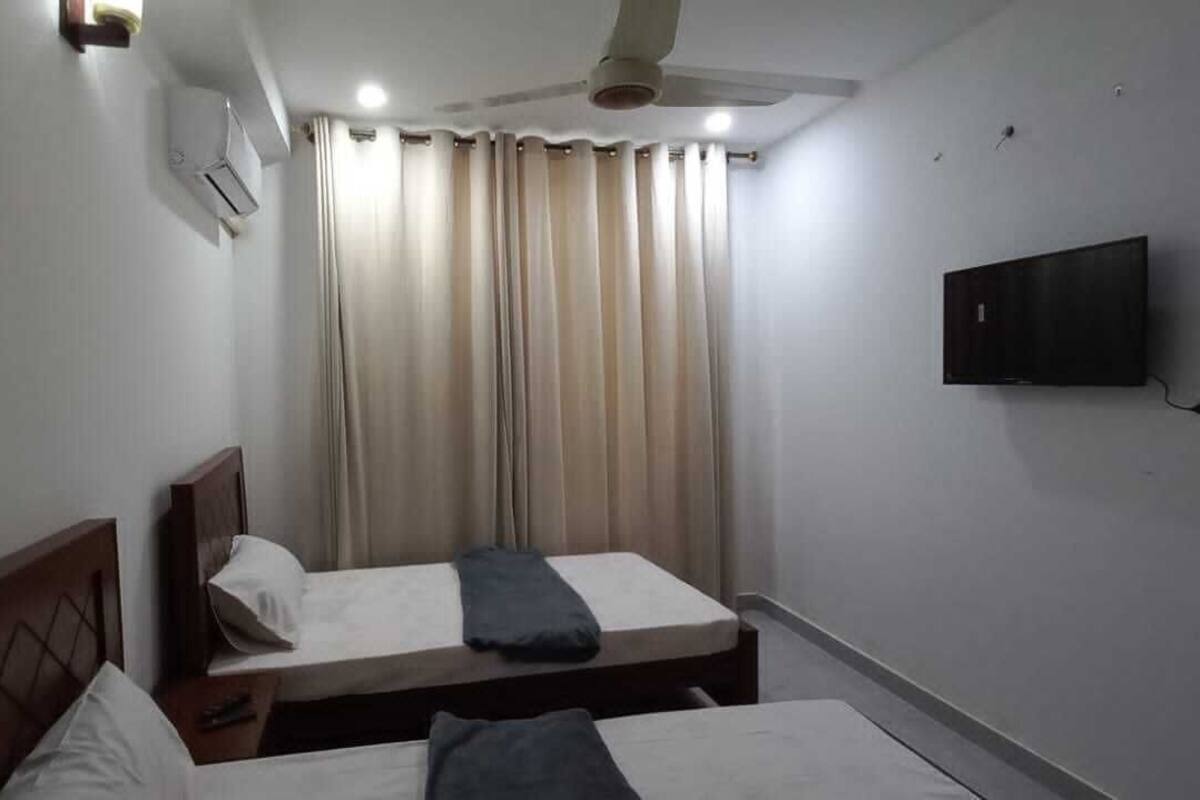 A hotel room with two single beds, a ceiling fan, an air conditioning unit, a TV mounted on the wall, and a window with curtains.