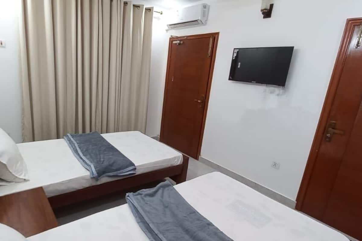A hotel room with two single beds, a wooden wardrobe, a television on the wall, and a window with curtains.