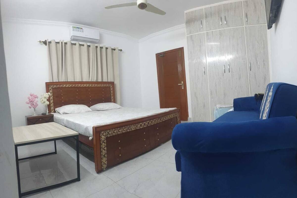 A bedroom with a bed, a blue sofa, a wooden wardrobe, a nightstand, and a ceiling fan.