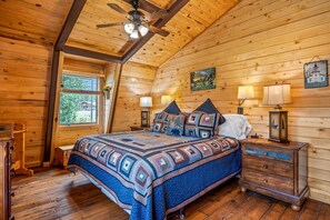 Een gezellige blokhut slaapkamer met een groot bed, houten wanden, een plafondventilator en een raam met uitzicht op groen.