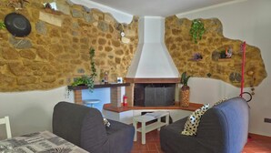 Uma sala de estar aconchegante com paredes de pedra, lareira, dois sofás cinzentos, uma mesa de jantar com toalha estampada e um pequeno bar.