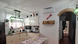 Uma cozinha compacta com armários brancos, uma mesa de jantar com toalha de mesa estampada e uma planta pendente.