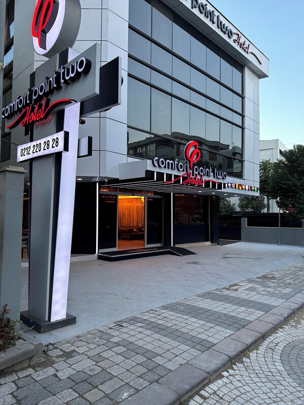 A modern hotel with a large glass facade, a prominent red and black logo, and a contact number displayed.