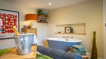 A modern bathroom with a freestanding bathtub, a large mirror, and a wooden shelf with books and a plant.