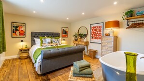 A bedroom with a large bed, a bedside table, a lamp, a mirror, a dresser, and a bathtub. - Haxby Hayloft: A Charming Countryside Retreat Near York (York)