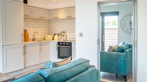 A modern kitchen with white cabinets, a built-in oven, and a sink. There is a teal sofa in the living area, and a large mirror on the wall. - Bootham Row 8: A Cozy City-Centre Retreat in York (York)