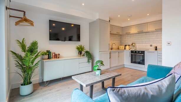 A modern kitchen with white cabinets, a built-in oven, and a countertop with a kettle. There is a dining area with a blue sofa and a small table. - Bootham Row 8: A Cozy City-Centre Retreat in York (York)