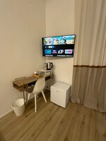 Una habitación pequeña y moderna con un escritorio de madera, una silla blanca, un televisor montado en la pared, una cafetera y un refrigerador pequeño.