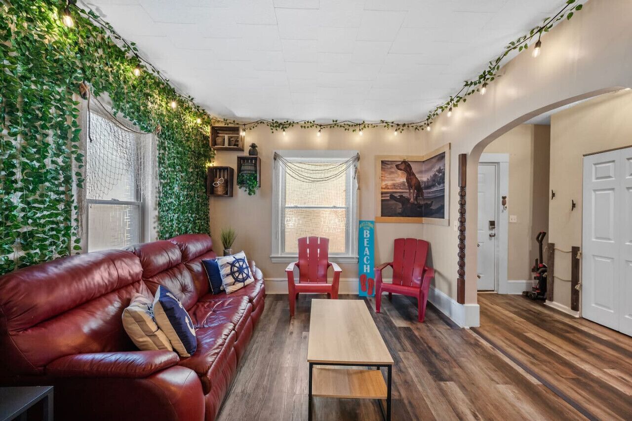 A cozy living room with a leather sofa, red chairs, a wooden coffee table, and a wall adorned with greenery and string lights.