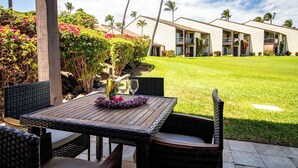 A patio with wicker furniture and a table set for two, overlooking a well-maintained lawn and a building with multiple balconies. - Hale Kamaole #124 Ground floor condo, easy access to best beach on Maui! (Kihei)