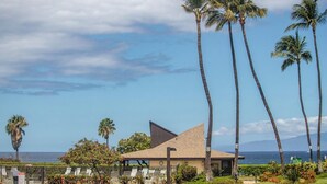 A modern building with a triangular roof, surrounded by palm trees and a view of the ocean. - Hale Kamaole #124 Ground floor condo, easy access to best beach on Maui! (Kihei)