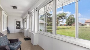 Varanda coberta com paredes brancas e piso de madeira, grandes janelas com vista para um gramado verde e edifícios distantes.