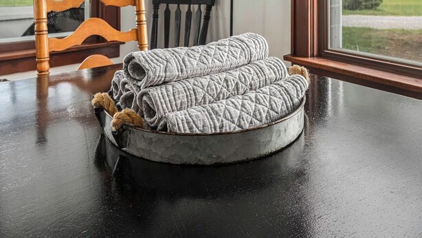 Uma cama redonda e texturizada para cachorro sobre uma mesa de madeira escura, com uma janela ao fundo.