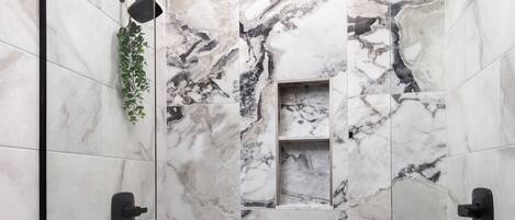 A modern shower with marble tiles, a glass door, and a small shelf with a plant.