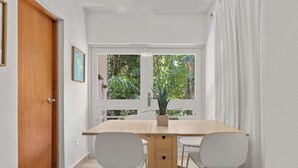Een eethoek met een houten tafel en twee witte stoelen, een bloempot op de tafel, en uitzicht op groen door een raam.