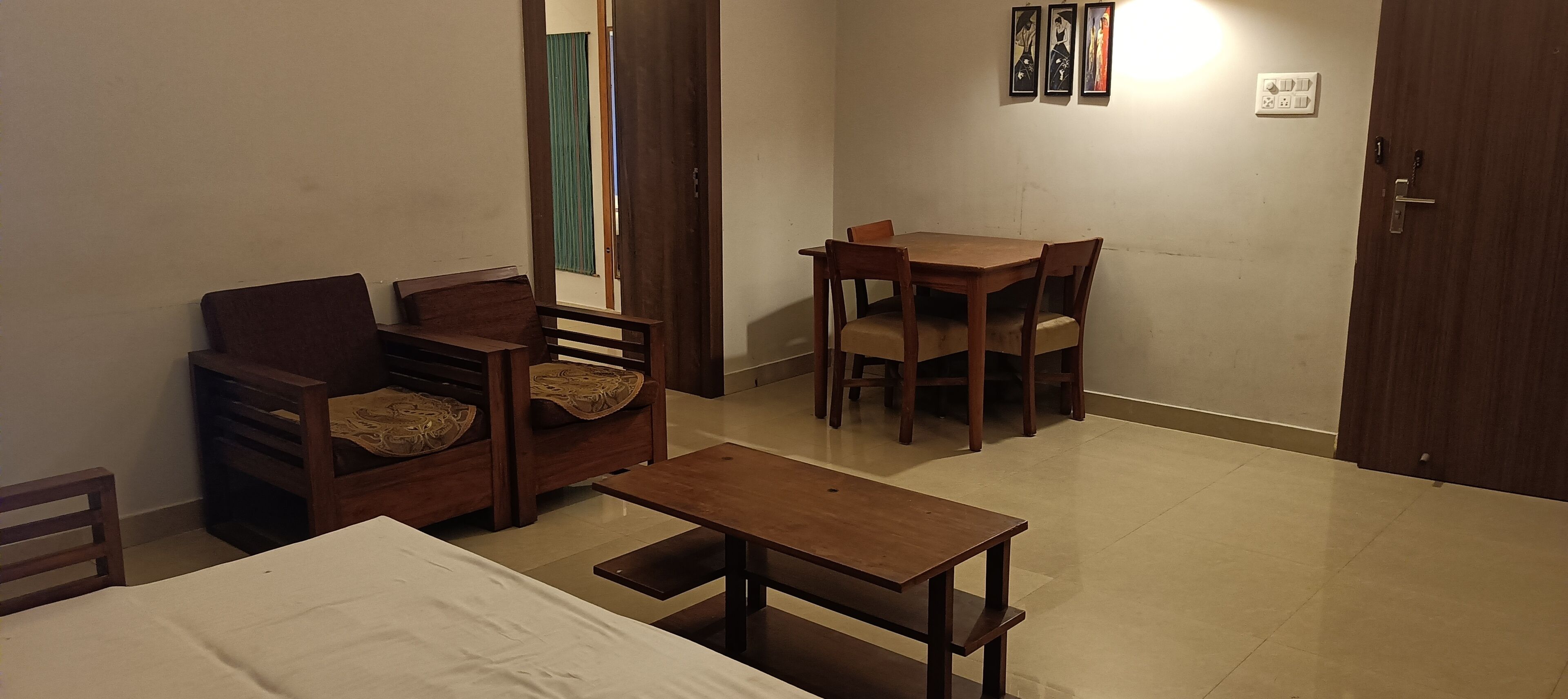 A small, well-lit room with a bed, a wooden table, two chairs, and a sofa. There is a door and a wall-mounted light fixture.