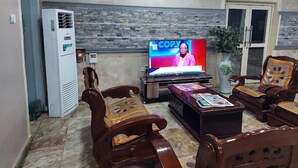 A living room with a television displaying a news program, a stone wall, a refrigerator, and wooden furniture. - Fingerprints Hotel (Oba Ile)