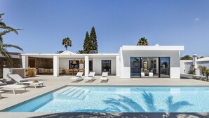 A modern poolside area with lounge chairs, a dining area, and a glass-enclosed room with a view of the pool. - Villa Bellagio - Rent Villas Lanzarote (Playa Blanca)