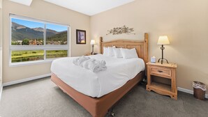 Una cama bien tendida con sábanas blancas y una toalla, una mesita de noche de madera con una lámpara, un cuadro en la pared y una ventana con vistas a la montaña.