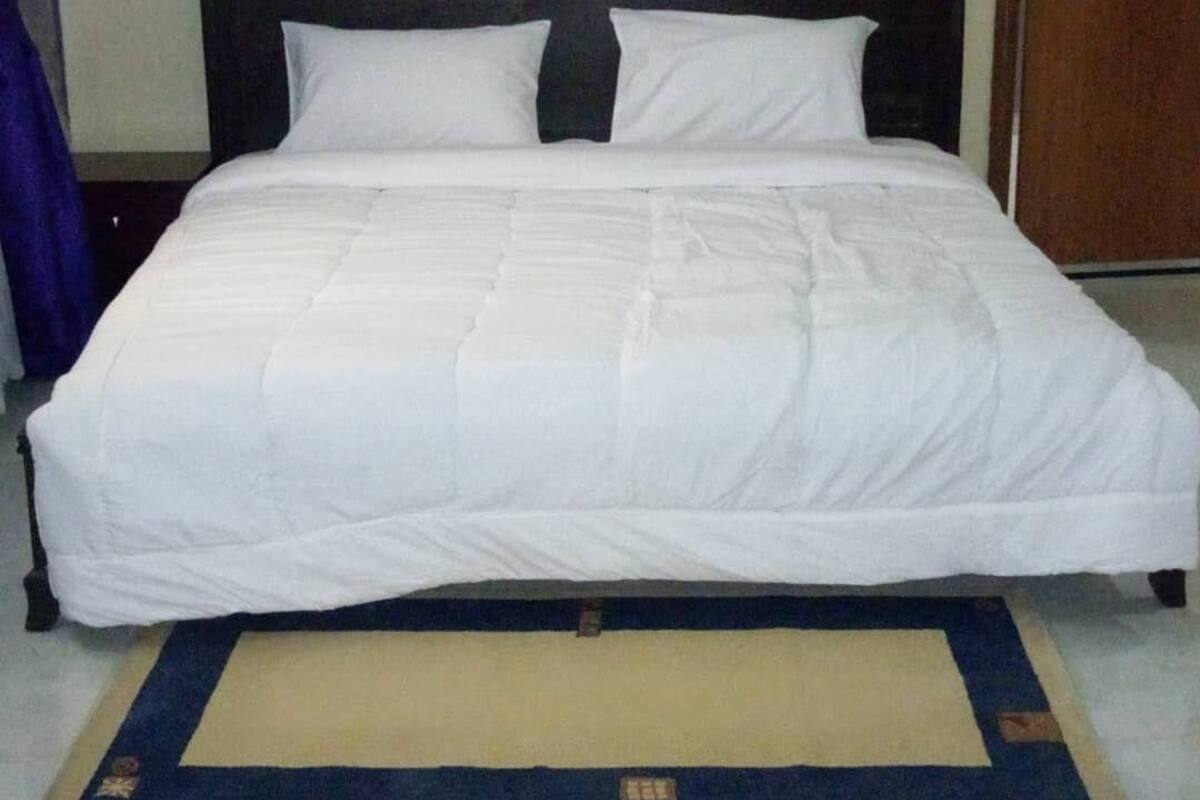 Una habitación de hotel estándar con una cama individual, cabecera de madera oscura, ropa de cama blanca y una alfombra beige con borde azul.