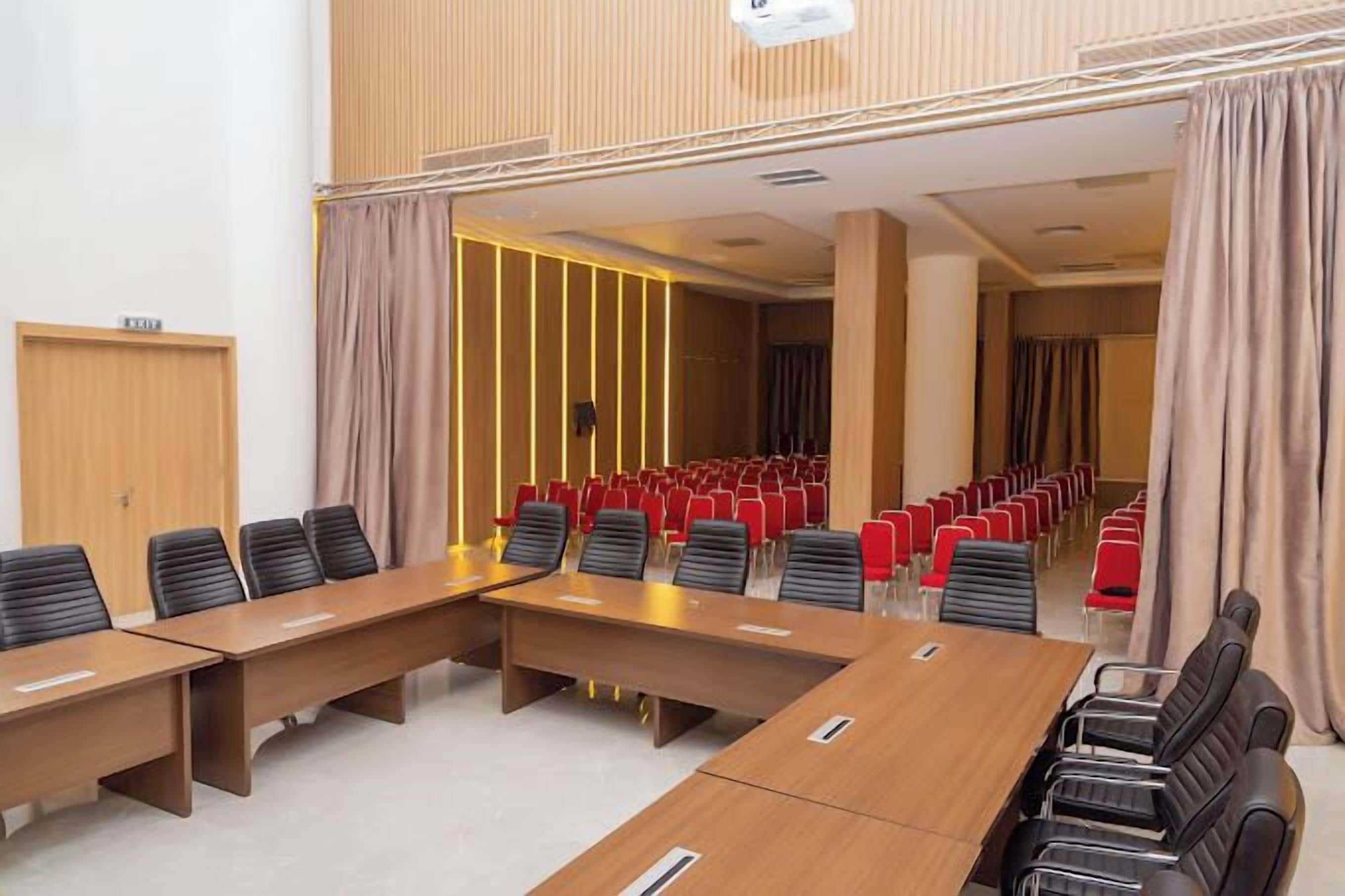 A conference room with long tables and chairs, a wooden panel wall, and a view into a corridor with red chairs.