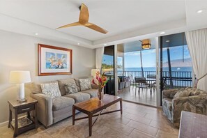 Sala de estar com sofá, mesa de centro e uma pintura na parede. Há uma varanda com vista para o oceano.
