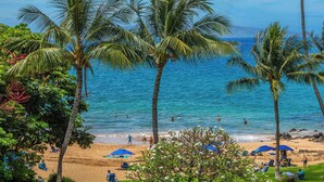 A beach with palm trees, people swimming, and umbrellas on the sand.