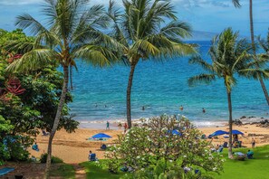 A beach with palm trees, people swimming, and umbrellas on the sand.