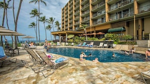 A hotel pool area with lounge chairs and umbrellas, surrounded by palm trees and a multi-story building.