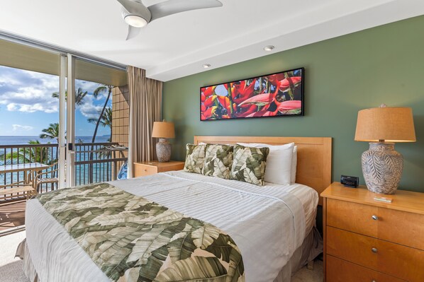 Una habitación de hotel con cama, dos mesitas de noche con lámparas, un ventanal con vistas al océano y un cuadro con motivos tropicales en la pared.
