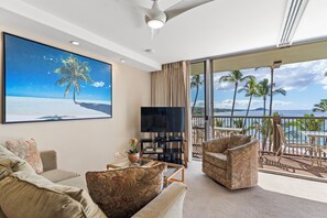 Uma sala de estar com uma TV grande, assentos confortáveis, uma estante e uma varanda com vista para palmeiras e o oceano.