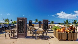 An outdoor seating area with tables and chairs, a grill station, and decorative planters. - Oceanfront Bliss at Royal Mauian #405–Stunning Views, Prime South Kihei Location (Kihei, Maui)