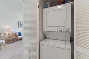 Pesuhuone, jossa on etuaukollinen pesukone ja kuivausrumpu, rottinkituoli, pieni pöytä ja lamppu.