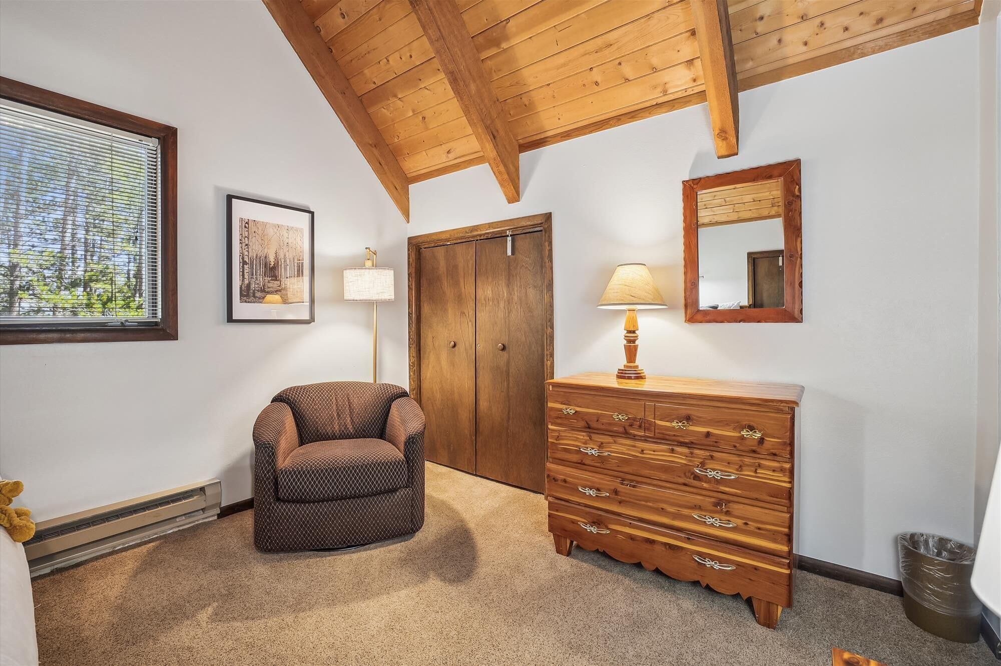 A room with a wooden ceiling, a wooden dresser with drawers, a chair, a framed picture on the wall, and a floor lamp.
