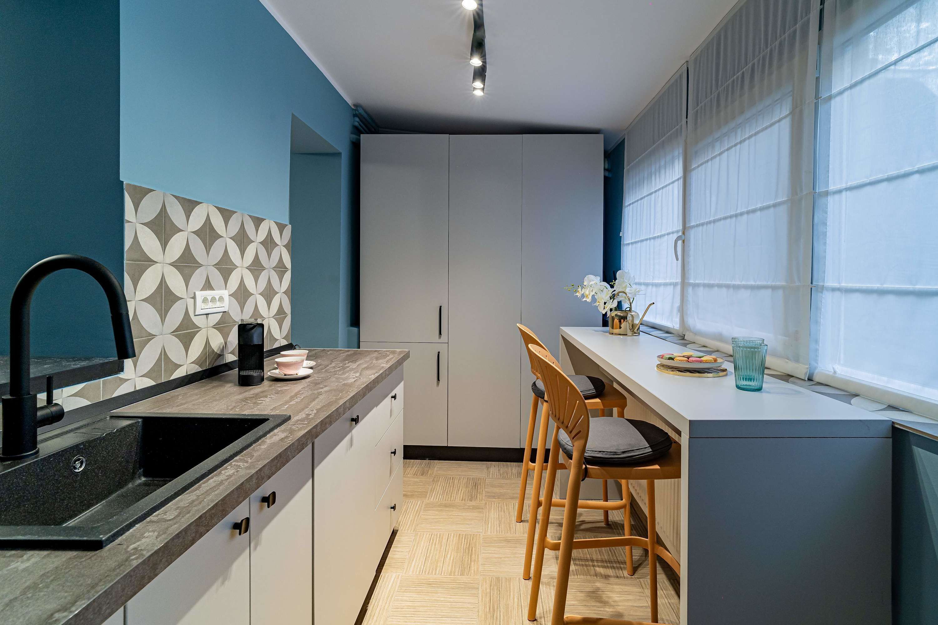 A modern kitchen with a black sink, white cabinets, and a patterned backsplash. There are two wooden chairs and a small table with a glass container.
