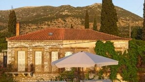 A stone building with a tiled roof, outdoor seating with umbrellas, and a mountainous backdrop.