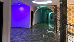 Un couloir d’hôtel moderne avec un sol en marbre, des luminaires verts et blancs au plafond et un mur orné d’un motif géométrique.