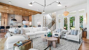 A spacious living area with a large white sofa, a coffee table, and a fireplace. There is a kitchen in the background with wooden cabinets and a staircase leading to an upper level. - Sleeps 18! Private Pool, Spa, LSV/Golf Cart! Gulf Views from Rooftop! Pet Friendly! (Santa Rosa Beach)