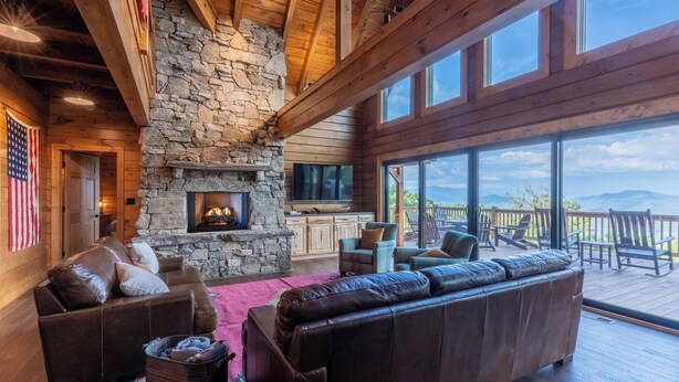 A cozy living room with a stone fireplace, leather sofa, and a large window overlooking a deck. - High Country Cabin -  Mountain & Fontana Lake Views - Hot Tub (Almond)