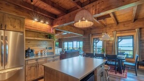 A spacious kitchen with wooden cabinets, a stainless steel refrigerator, and a large island. There is a dining area with a table and chairs, and large windows providing a view of the outdoors. - High Country Cabin -  Mountain & Fontana Lake Views - Hot Tub (Almond)