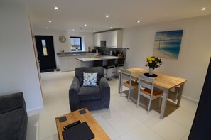 A modern living room with a grey sofa, a wooden coffee table, and a dining area with a wooden table and white chairs. A kitchen with white cabinets and a window is visible in the background.