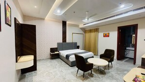 A hotel room with a bed, two chairs, a small table, a nightstand, a desk, a mirror, a window with curtains, and a ceiling fan. - Hotel Miracle Resorts (Phagwara)