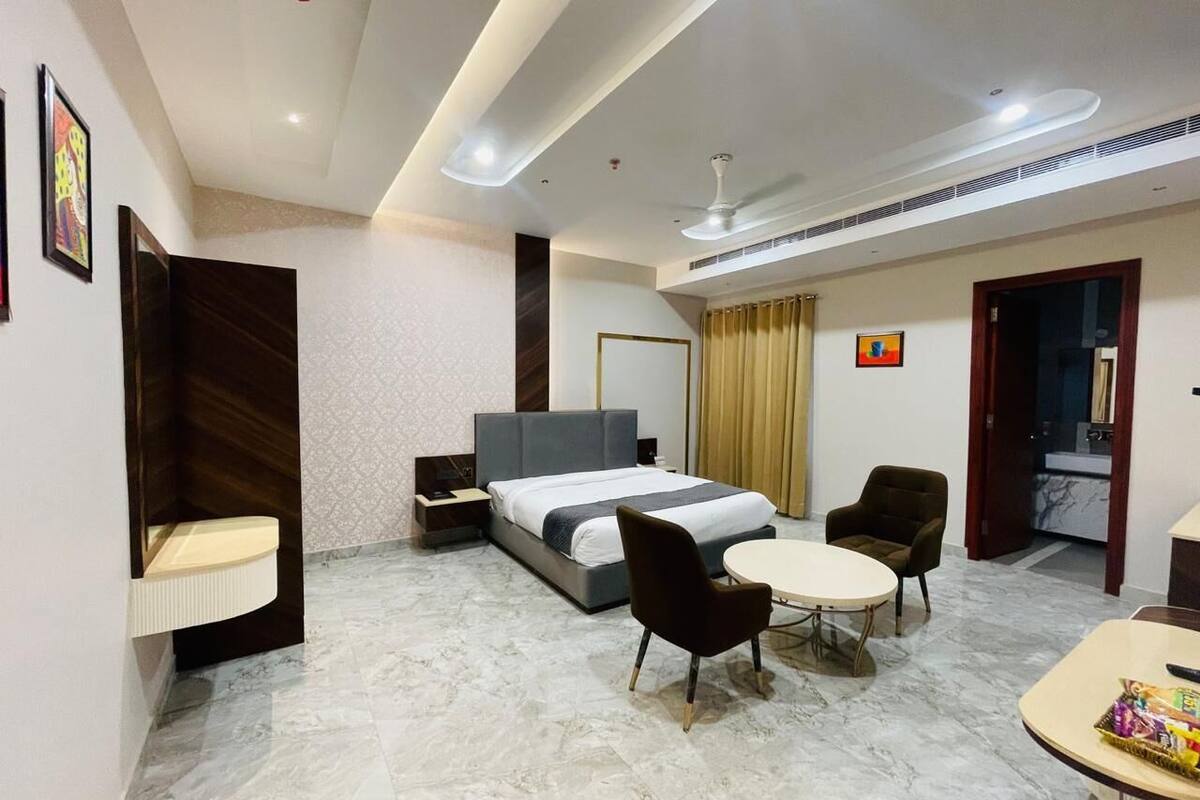 A hotel room with a bed, two chairs, a small table, a nightstand, a desk, a mirror, a window with curtains, and a ceiling fan.