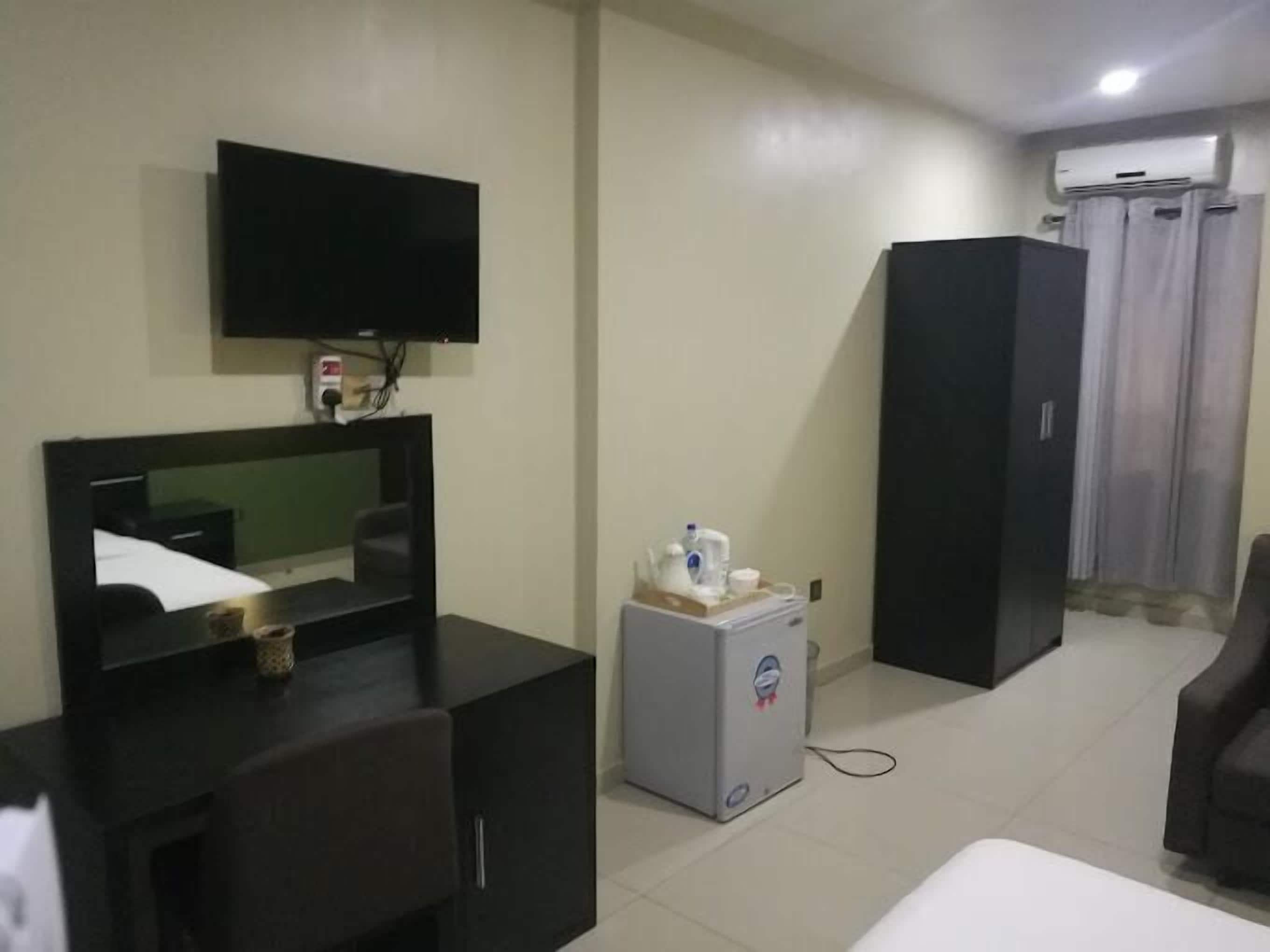 A hotel room with a flat-screen TV mounted on the wall, a small refrigerator, a dark-colored wardrobe, and a bed with a white mattress.