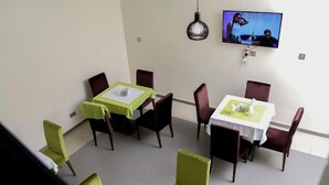 Zona pranzo con due tavoli, tovaglie verdi e bianche e sedie abbinate. Un televisore è montato a parete.