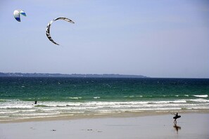 A beach with waves, a person walking, and a kite in the sky.