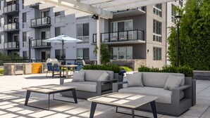 Un espace extérieur aménagé avec un canapé et des tables, un barbecue et une terrasse offrant une vue sur un bâtiment moderne.