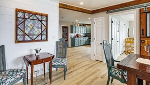 A dining area with a wooden table and chairs, a stained glass window, and a kitchen in the background. - Family Beach Days: Dog-friendly Cape Cod Condo (Dennis Port)